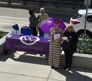 Ordain Women executive board members standing by a table on the sidewalk. The table has a purple Ordain Women cover.