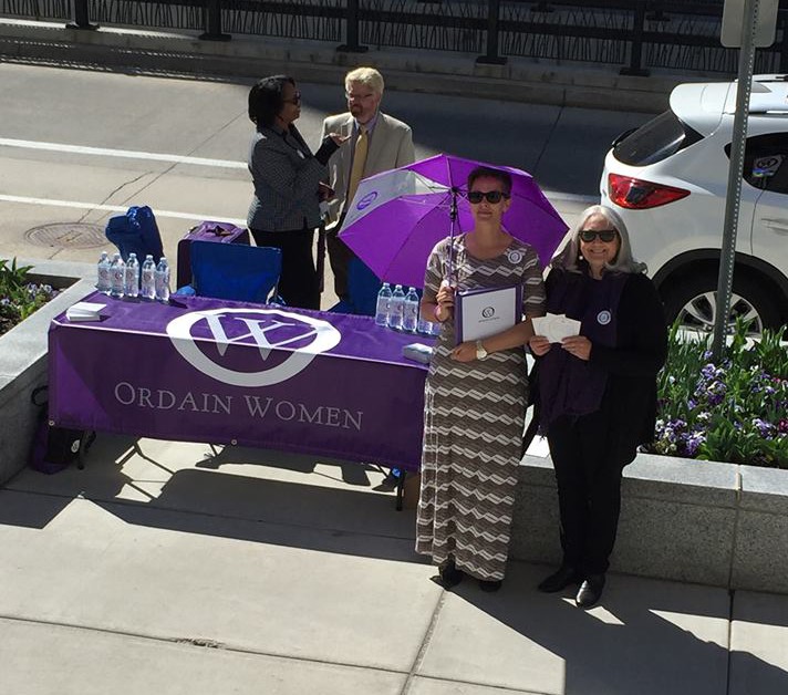 Members of Ordain Women's executive board are standing on a sidewalk, around a table that is covered with an Ordain Women table cloth. 