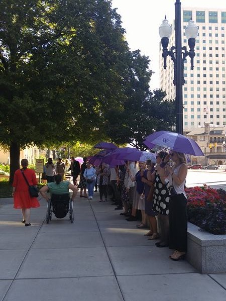 The sidewalk in front of the LDS Church Administration Building in Salt Lake City, UT is filled with people. Along the side is a line of men and women holding purple Ordain Women umbrellas for the Let My Voice Be Heard action.
