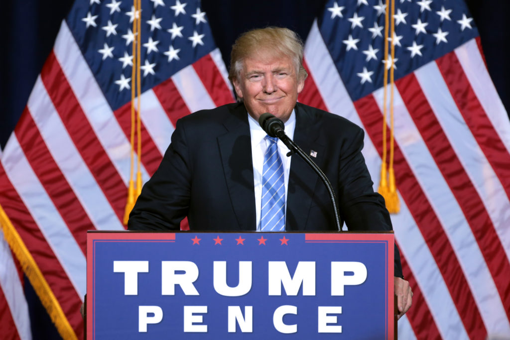 A picture of Donald J. Trump standing in front of two American flags and behing a podium covered in a sign that says, "Trump, Pence."