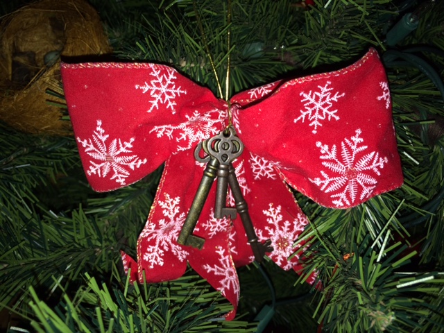 A picture of an ornament hanging from a tree. The ornament is a red Christmas bow, with white snowflakes on the fabric. There are three old-fashion keys hanging from the knot of the bow.