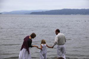 A man and a woman lead a young girl into the ocean. The man and girl are wearing white baptismal clothes.