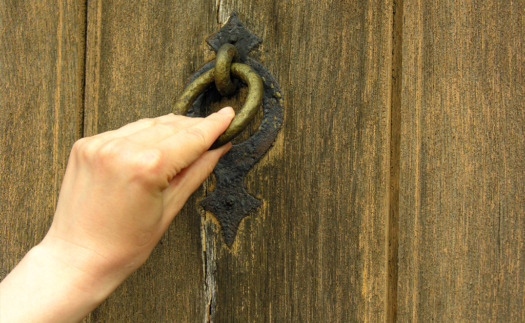 A picture of a wooden door, a hand is knocking on the door using the knocker.