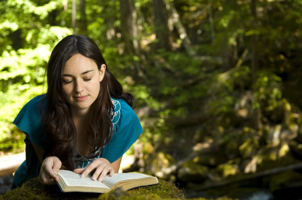 Woman kneeling at a rock in the woods, reading scriptures that are laying open on the rock.